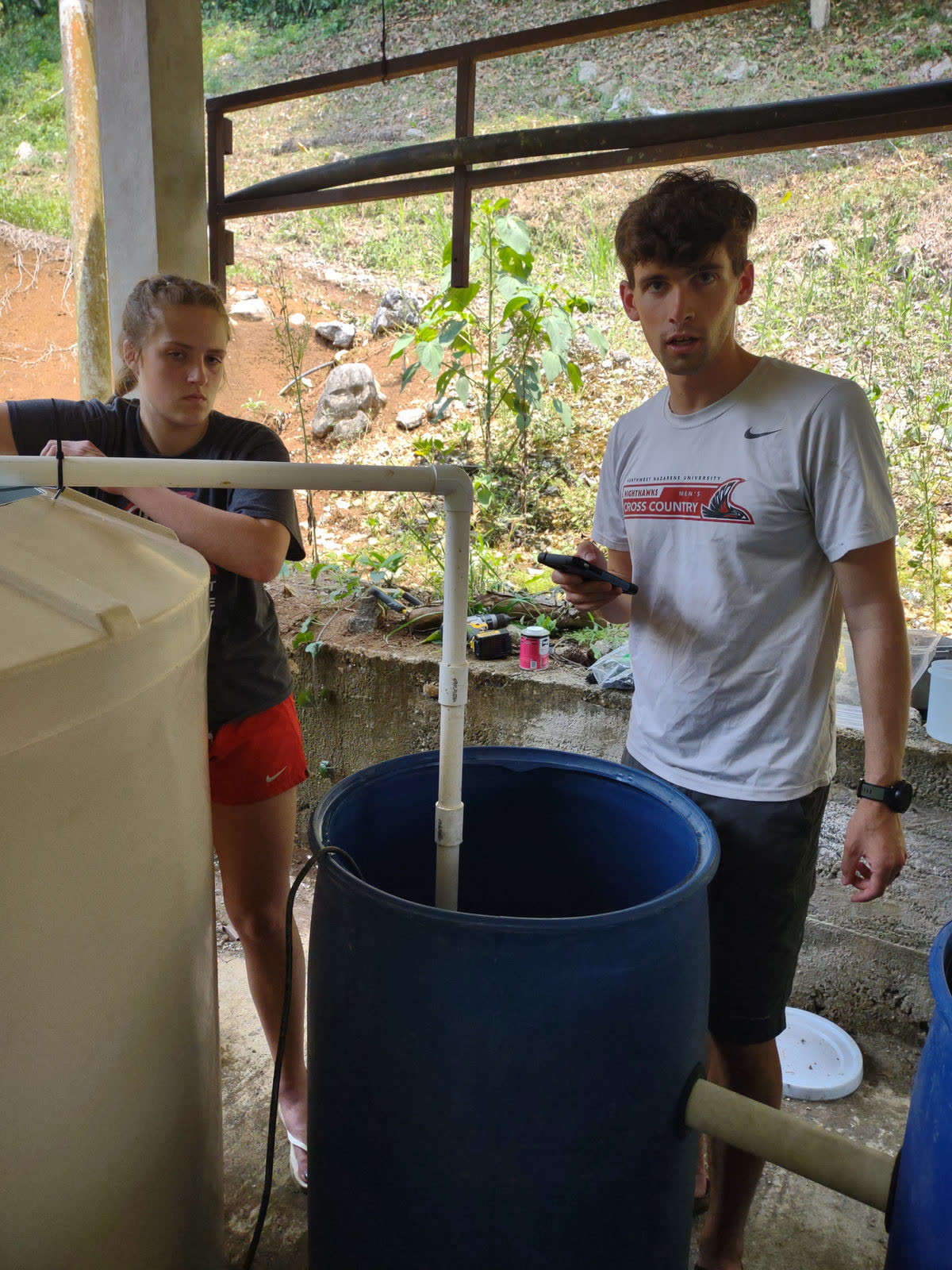 Two young men fill a blue barrel from a water filtration tank