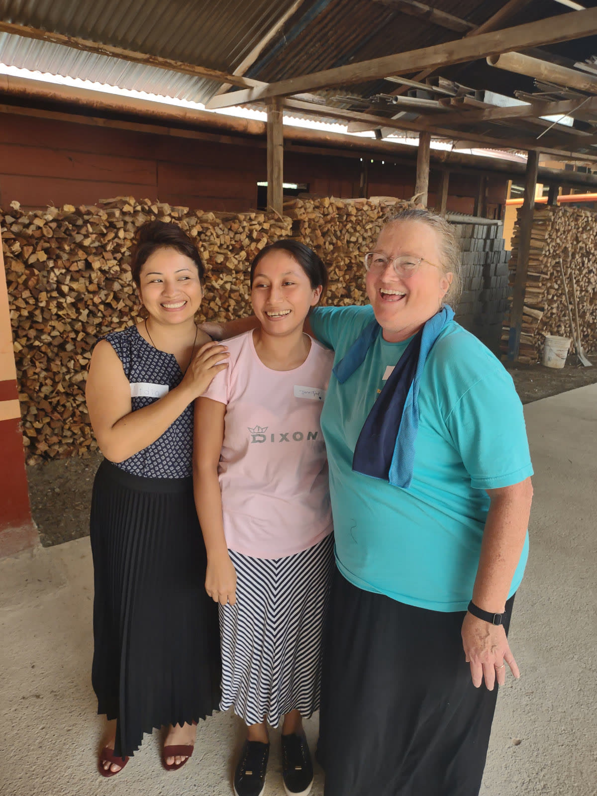 Three women smile in front of stacked firewood