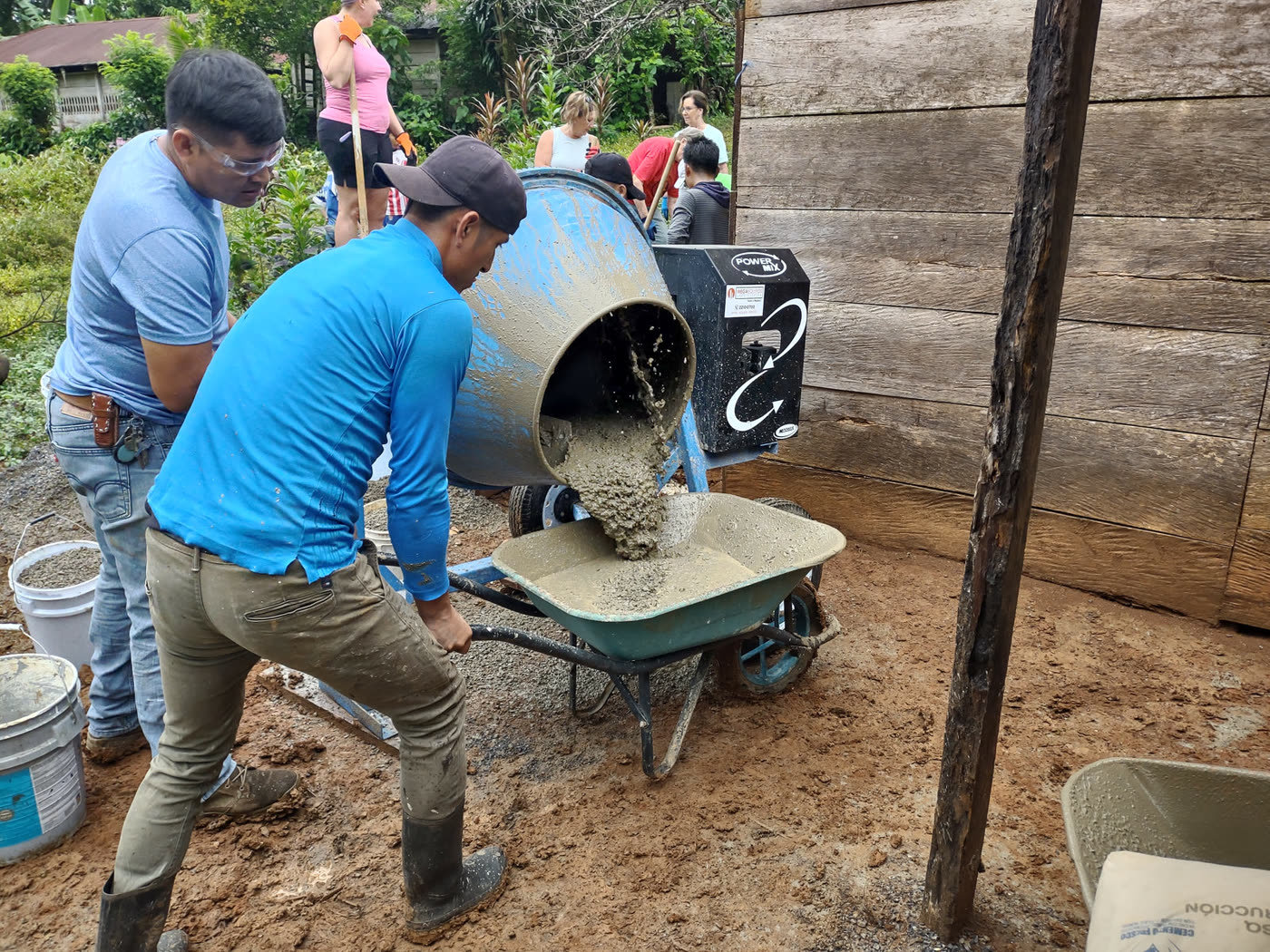 Two men pour concrete from a mixer into a wheelbarrow beside a wooden home